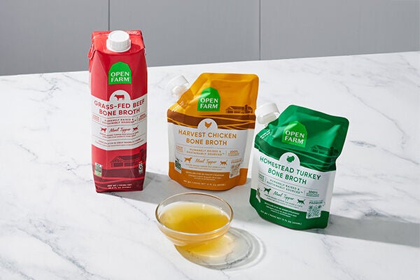 Three containers of Open Farm bone broth, one red, one orange, one green behind a clear bowl filled with broth on a white marble counter. 